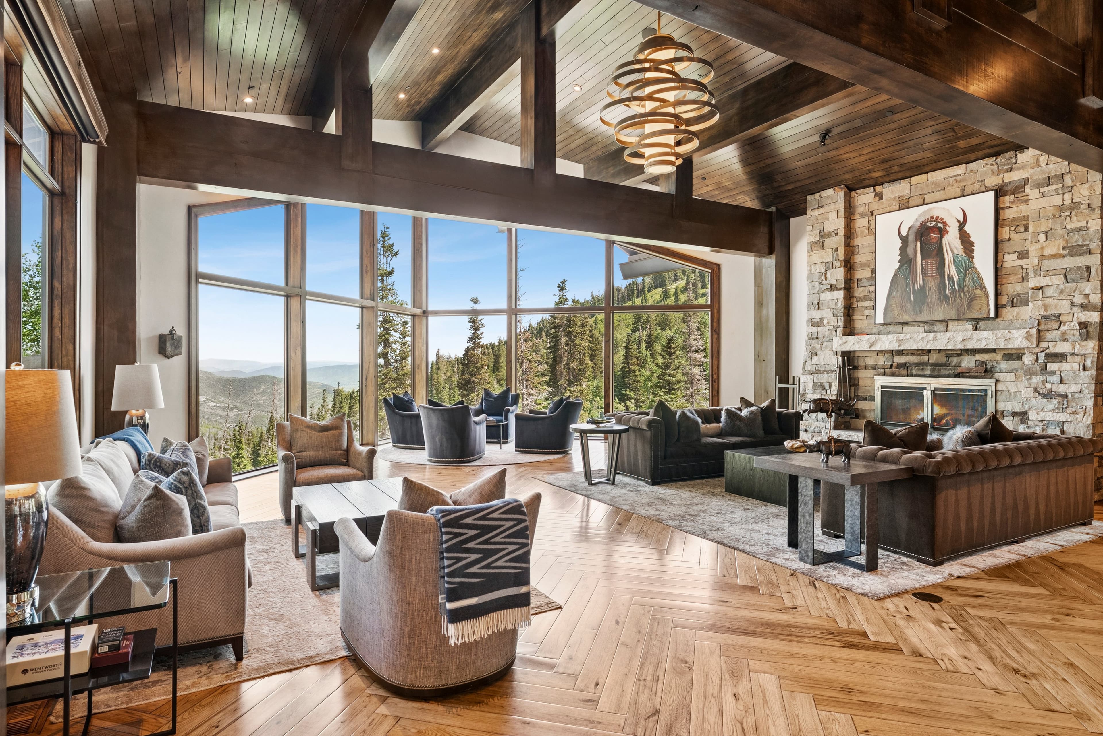 view of the grand room with two walls of windows overlooking the forest and mountains of park city. there is luxury furnishings and designer lighting and tall ceilings with giant wood beams